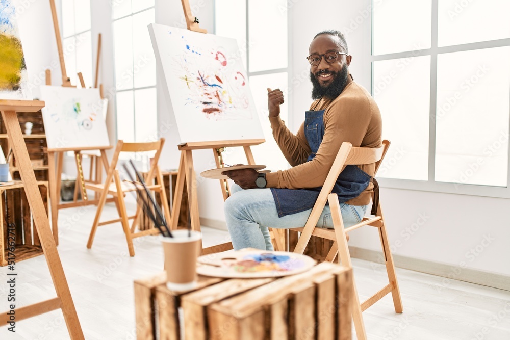 African american artist man painting on canvas at art studio pointing thumb up to the side smiling happy with open mouth