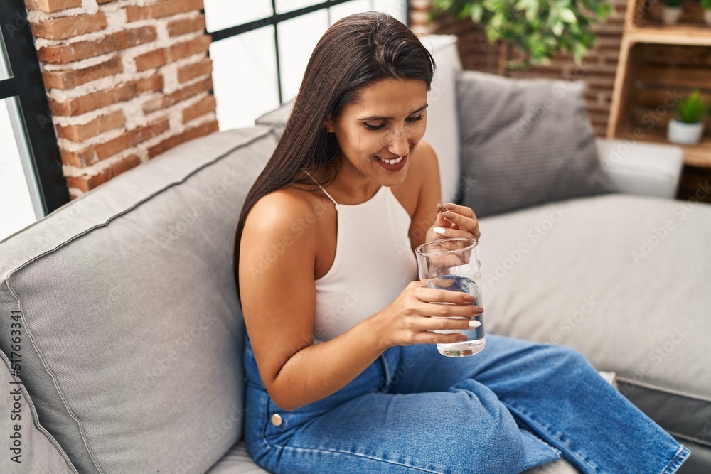 Young hispanic woman taking pill drinking water sitting on sofa at home Young hispanic woman taking pill drinking water sitting on sofa at home