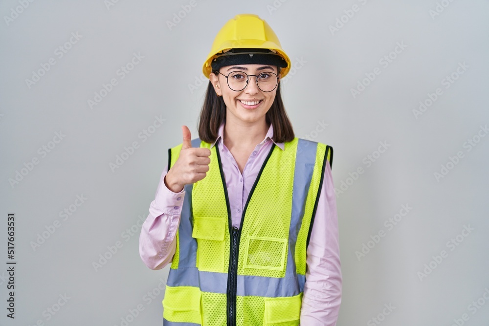 Hispanic girl wearing builder uniform and hardhat doing happy thumbs up gesture with hand. approving expression looking at the camera showing success. Hispanic girl wearing builder uniform and hardhat doing happy thumbs up gesture with hand. approving expression looking at the camera showing success.
