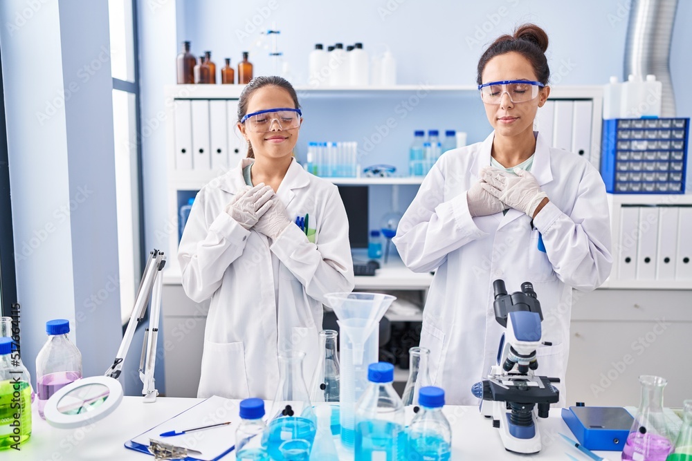 Young mother and daughter at scientist laboratory smiling with hands on chest with closed eyes and grateful gesture on face. health concept. Young mother and daughter at scientist laboratory smiling with hands on chest with closed eyes and grateful gesture on face. health concept.