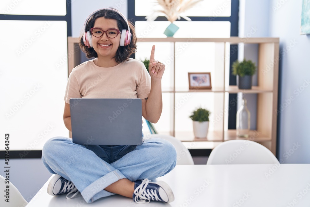 Young hispanic woman using laptop sitting on the table wearing headphones showing and pointing up with finger number one while smiling confident and happy. Young hispanic woman using laptop sitting on the table wearing headphones showing and pointing up with finger number one while smiling confident and happy.