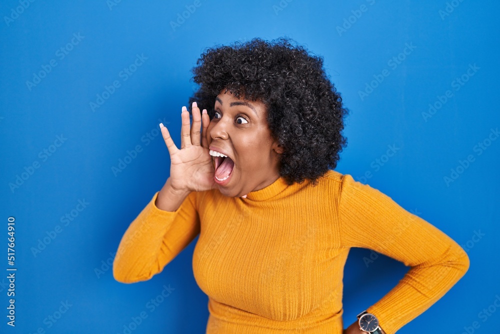 Black woman with curly hair standing over blue background shouting and screaming loud to side with hand on mouth. communication concept. Black woman with curly hair standing over blue background shouting and screaming loud to side with hand on mouth. communication concept.
