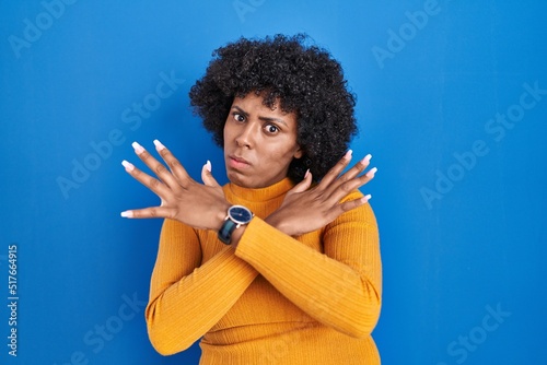 Black woman with curly hair standing over blue background rejection expression crossing arms doing negative sign, angry face