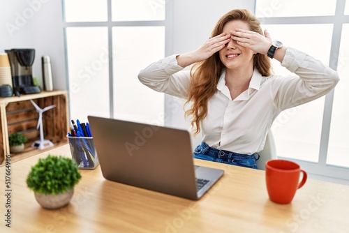 Young caucasian woman working at the office using computer laptop covering eyes with hands smiling cheerful and funny. blind concept.