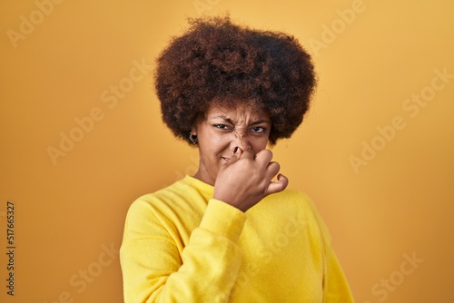 Young african american woman standing over yellow background smelling something stinky and disgusting, intolerable smell, holding breath with fingers on nose. bad smell © Krakenimages.com