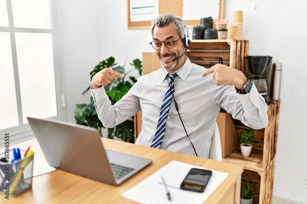 Middle age hispanic business man working at the office wearing operator headset looking confident with smile on face, pointing oneself with fingers proud and happy.