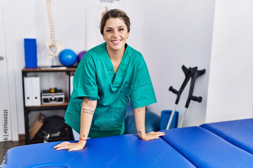 Young hispanic woman wearing physiotherapist uniform leaning on massage table at physiotherapy clinic