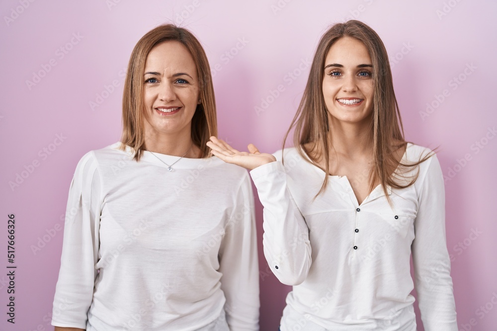 Middle age mother and young daughter standing over pink background smiling cheerful presenting and pointing with palm of hand looking at the camera. Middle age mother and young daughter standing over pink background smiling cheerful presenting and pointing with palm of hand looking at the camera.