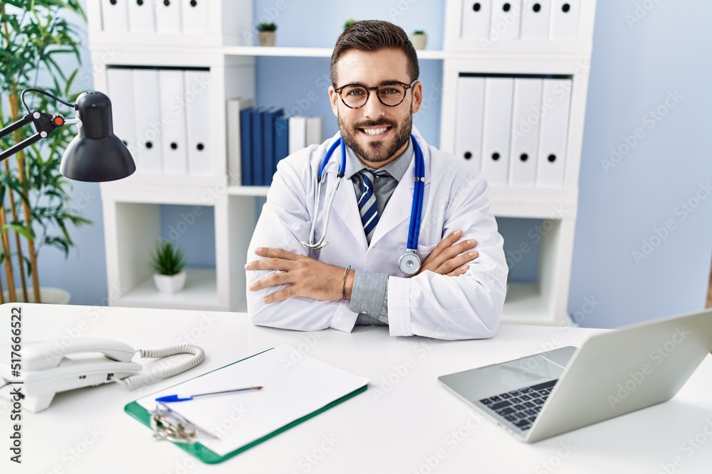 Obraz premium Young hispanic man wearing doctor uniform using laptop sitting at clinic