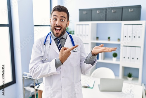 Handsome hispanic man wearing doctor uniform and stethoscope at medical clinic amazed and smiling to the camera while presenting with hand and pointing with finger.