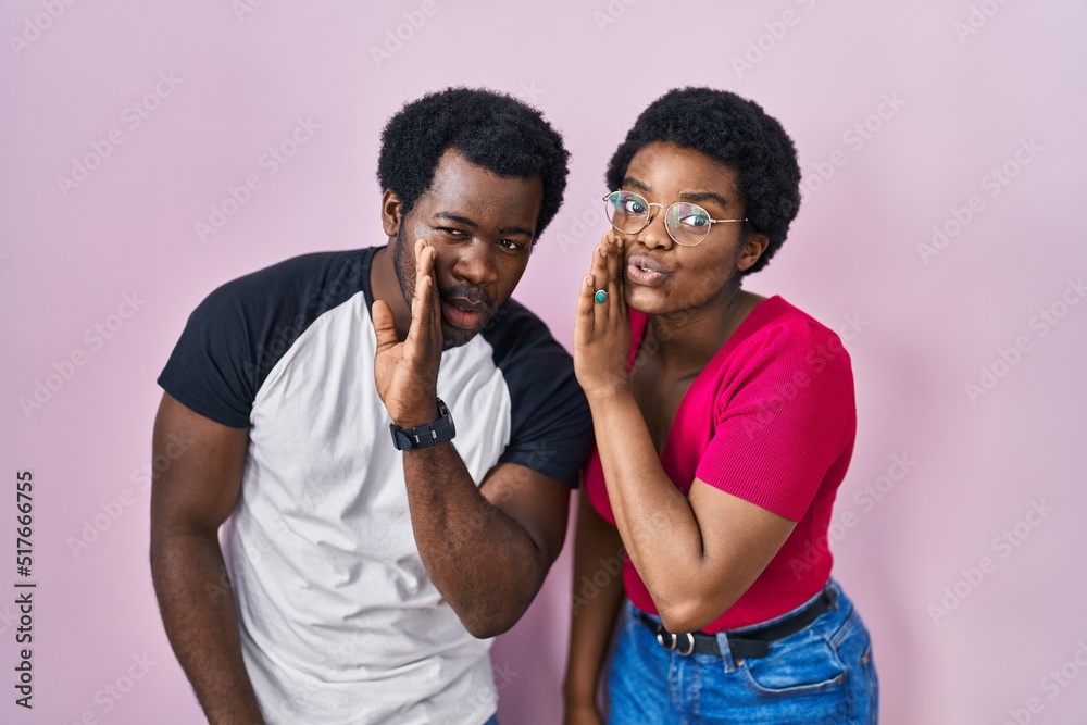Young african american couple standing over pink background hand on mouth telling secret rumor, whispering malicious talk conversation Young african american couple standing over pink background hand on mouth telling secret rumor, whispering malicious talk conversation