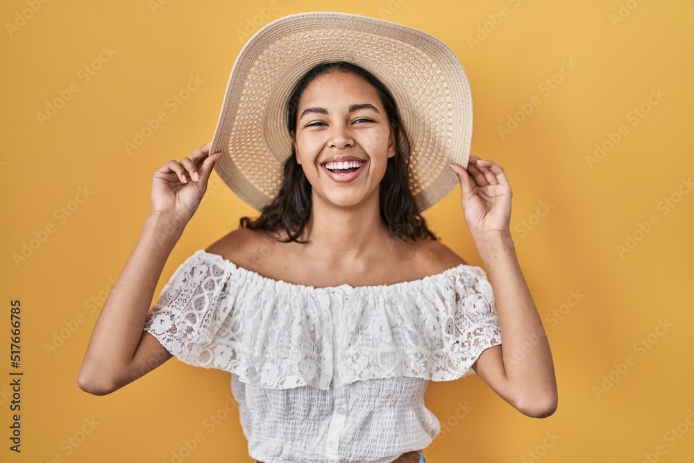 Young brazilian woman wearing summer hat over yellow background smiling and laughing hard out loud because funny crazy joke. Young brazilian woman wearing summer hat over yellow background smiling and laughing hard out loud because funny crazy joke.