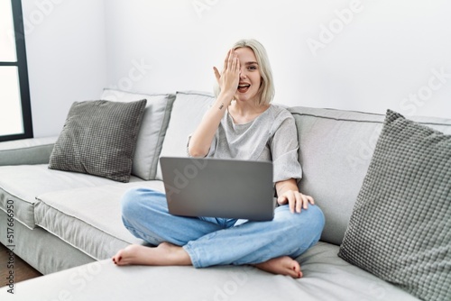 Young caucasian woman using laptop at home sitting on the sofa covering one eye with hand, confident smile on face and surprise emotion.