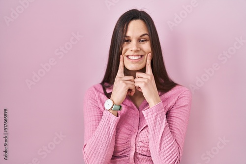 Young hispanic woman standing over pink background smiling with open mouth, fingers pointing and forcing cheerful smile