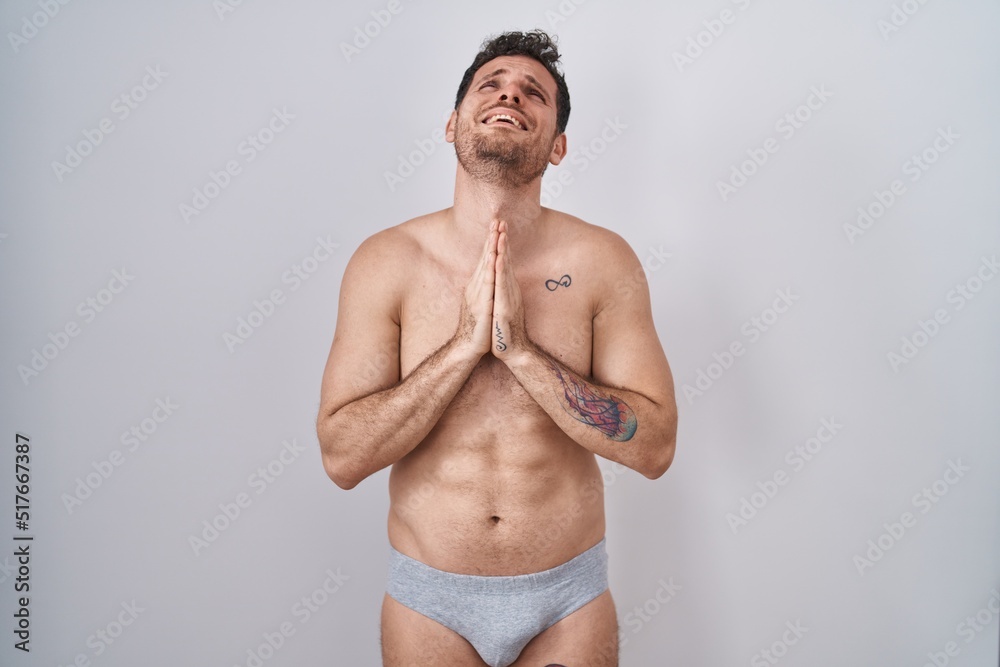 Fototapeta premium Young hispanic man standing shirtless wearing underware begging and praying with hands together with hope expression on face very emotional and worried. begging.