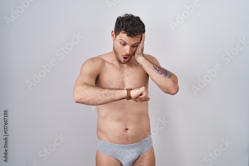 Young hispanic man standing shirtless wearing underware looking at the watch time worried, afraid of getting late