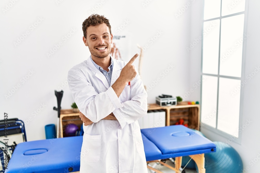 Handsome young man working at pain recovery clinic with a big smile on face, pointing with hand and finger to the side looking at the camera. Handsome young man working at pain recovery clinic with a big smile on face, pointing with hand and finger to the side looking at the camera.