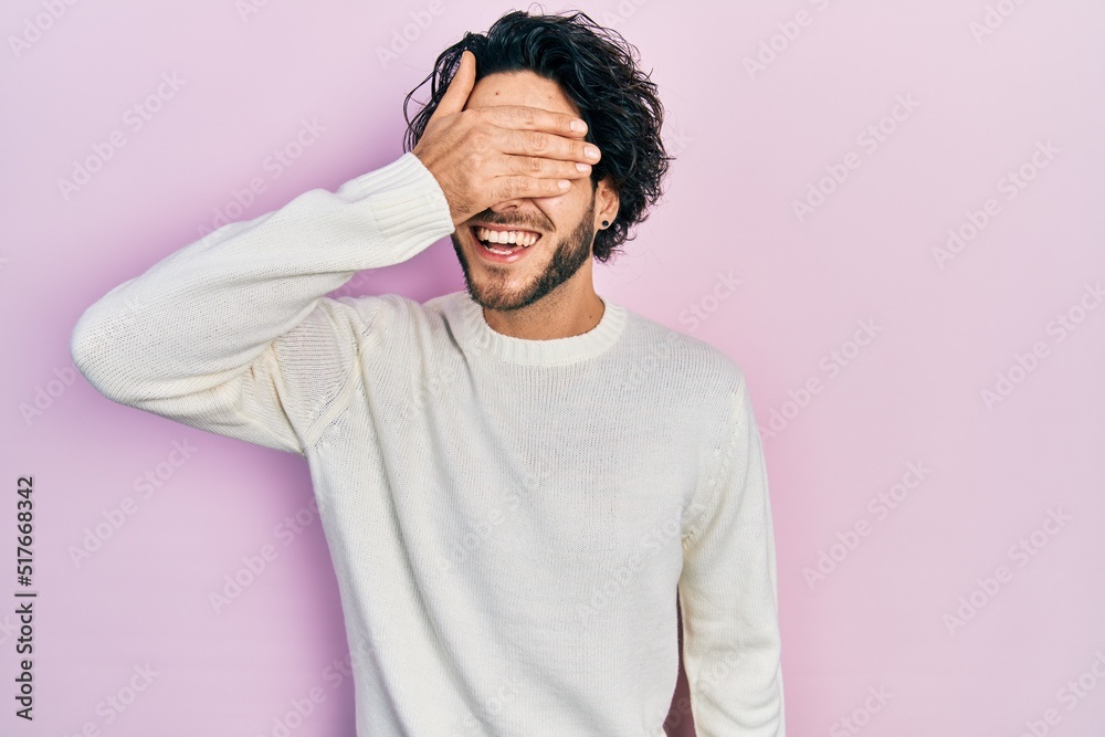Handsome hispanic man wearing casual white sweater smiling and laughing with hand on face covering eyes for surprise. blind concept.