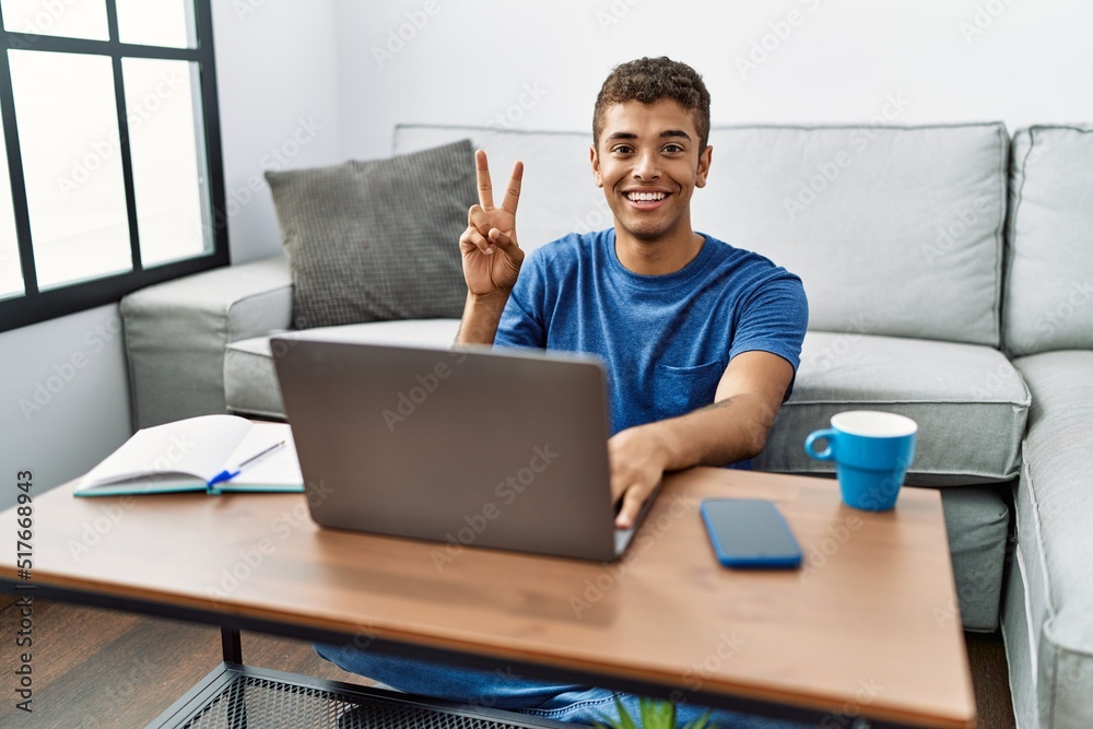 Young handsome hispanic man using laptop sitting on the floor showing and pointing up with fingers number two while smiling confident and happy. Young handsome hispanic man using laptop sitting on the floor showing and pointing up with fingers number two while smiling confident and happy.