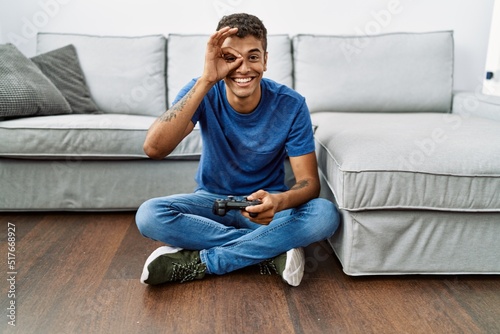 Young handsome hispanic man playing video game sitting by the sofa smiling happy doing ok sign with hand on eye looking through fingers © Krakenimages.com