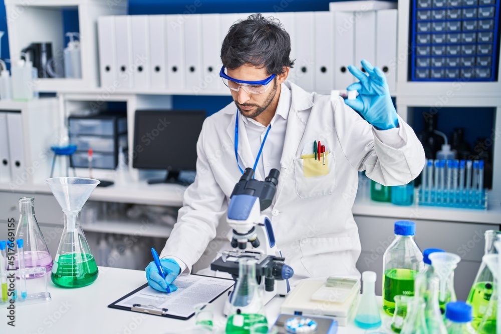Young hispanic man scientist write on clipboard holding sample at laboratory