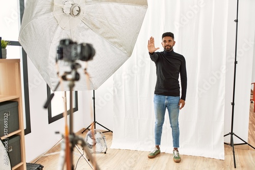 Young hispanic man with beard posing as model at photography studio doing stop sing with palm of the hand. warning expression with negative and serious gesture on the face.