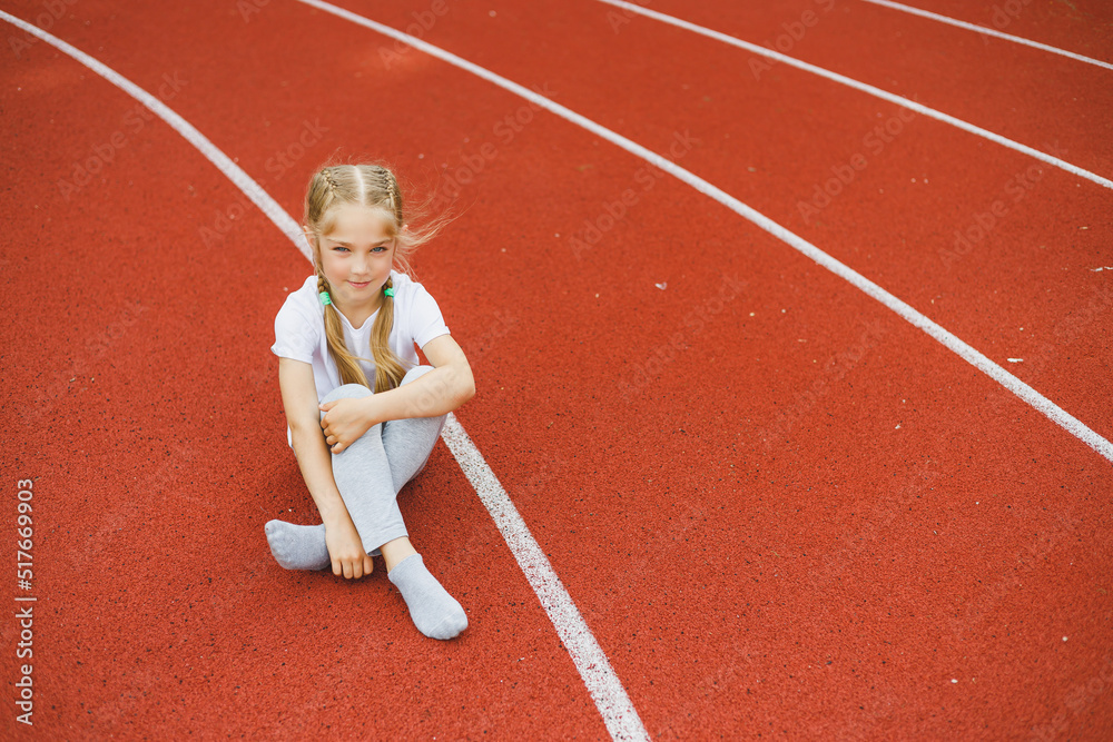 A little girl of 7-8 years old in a white t-shirt runs outdoors in the stadium. The girl is doing sports on a sunny day