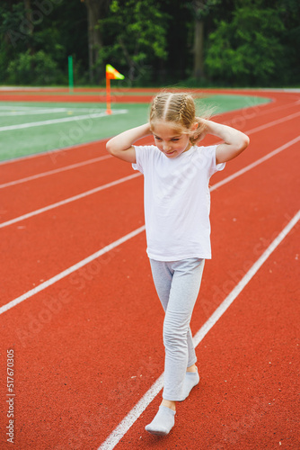 A little girl of 7-8 years old in a white t-shirt runs outdoors in the stadium. The girl is doing sports on a sunny day