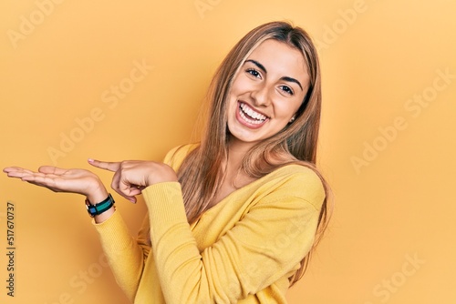 Beautiful hispanic woman wearing casual yellow sweater amazed and smiling to the camera while presenting with hand and pointing with finger.