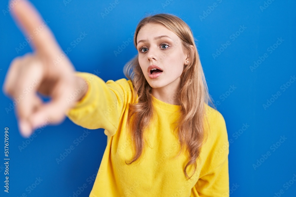 Young caucasian woman standing over blue background pointing with finger surprised ahead, open mouth amazed expression, something on the front Young caucasian woman standing over blue background pointing with finger surprised ahead, open mouth amazed expression, something on the front