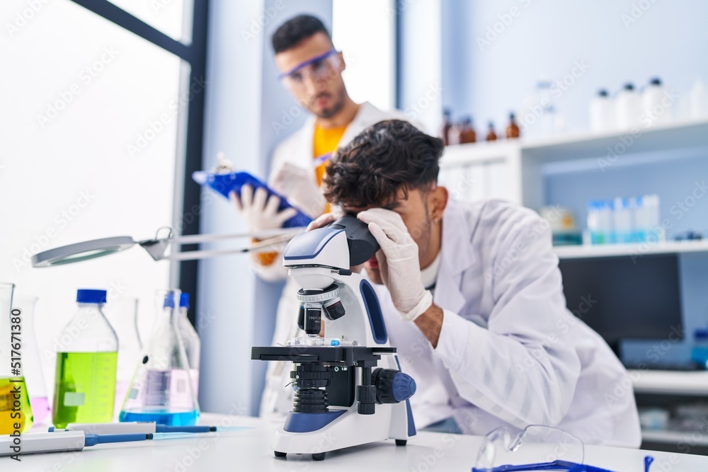 Fototapeta premium Two man scientists using microscope write on clipboard working at laboratory