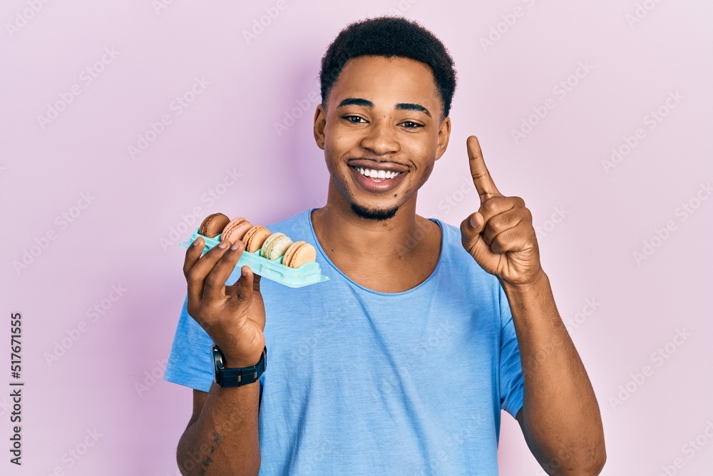 Young african american man holding macarons smiling with an idea or question pointing finger with happy face, number one Young african american man holding macarons smiling with an idea or question pointing finger with happy face, number one