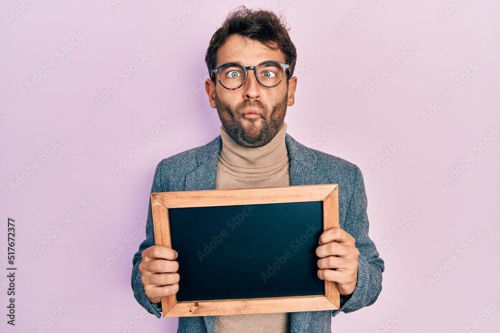 Handsome man with beard holding blackboard making fish face with mouth and squinting eyes, crazy and comical. Handsome man with beard holding blackboard making fish face with mouth and squinting eyes, crazy and comical.