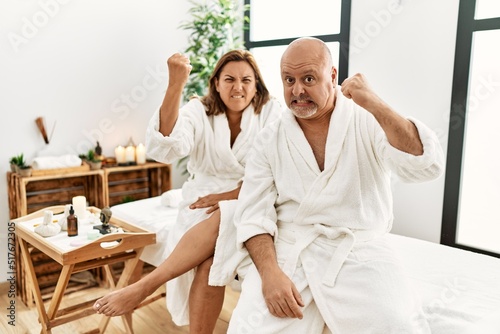 Middle age hispanic couple wearing bathrobe at wellness spa angry and mad raising fist frustrated and furious while shouting with anger. rage and aggressive concept.