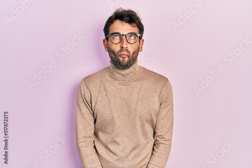 Handsome man with beard wearing turtleneck sweater and glasses making fish face with lips, crazy and comical gesture. funny expression.