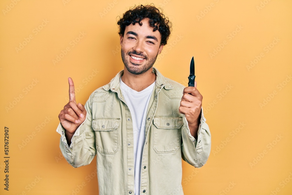 Young hispanic man holding pocket knife surprised with an idea or question pointing finger with happy face, number one