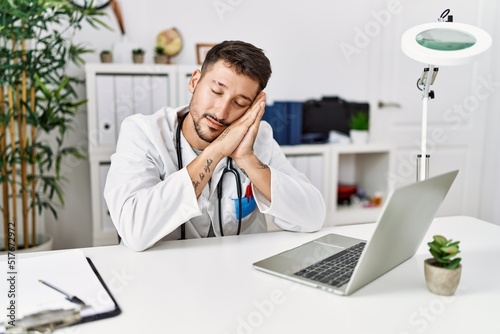 Young doctor working at the clinic using computer laptop sleeping tired dreaming and posing with hands together while smiling with closed eyes.