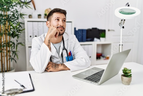 Young doctor working at the clinic using computer laptop with hand on chin thinking about question, pensive expression. smiling with thoughtful face. doubt concept.