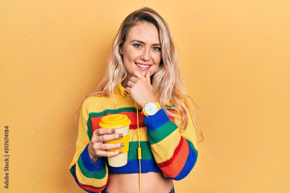 Beautiful young blonde woman drinking cup of coffee wearing headphones looking confident at the camera smiling with crossed arms and hand raised on chin. thinking positive. Beautiful young blonde woman drinking cup of coffee wearing headphones looking confident at the camera smiling with crossed arms and hand raised on chin. thinking positive.
