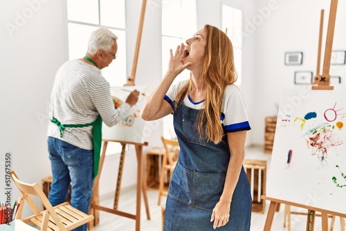 Hispanic woman wearing apron at art studio shouting and screaming loud to side with hand on mouth. communication concept.