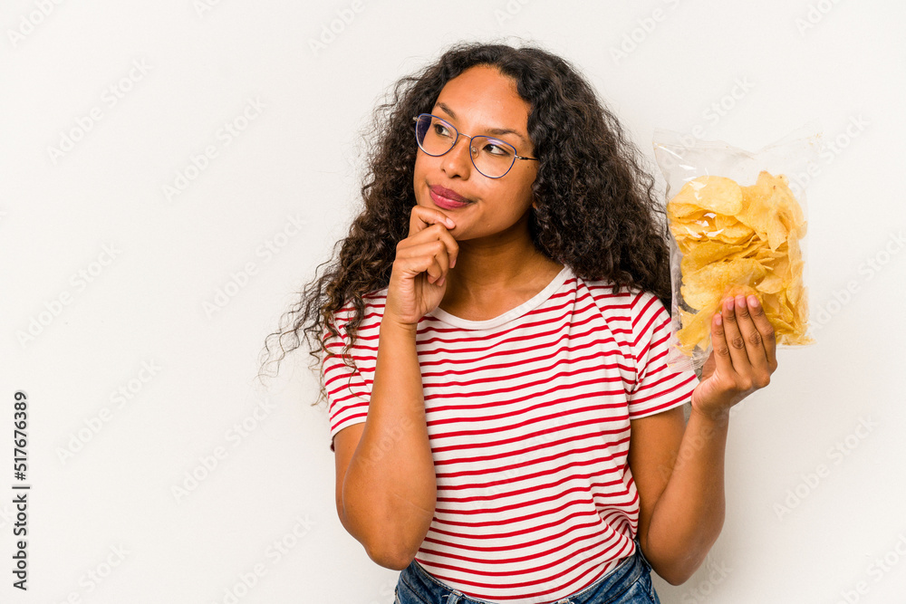 Young hispanic woman holding crips isolated on white background looking sideways with doubtful and skeptical expression. Young hispanic woman holding crips isolated on white background looking sideways with doubtful and skeptical expression.