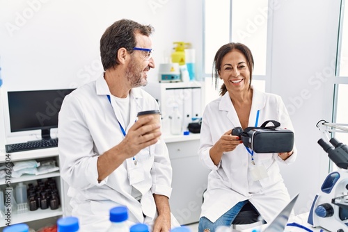 Middle age man and woman partners wearing scientist uniform drinking coffee and holding vr goggles at laboratory