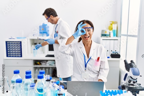 Middle age woman working at scientist laboratory smiling happy doing ok sign with hand on eye looking through fingers