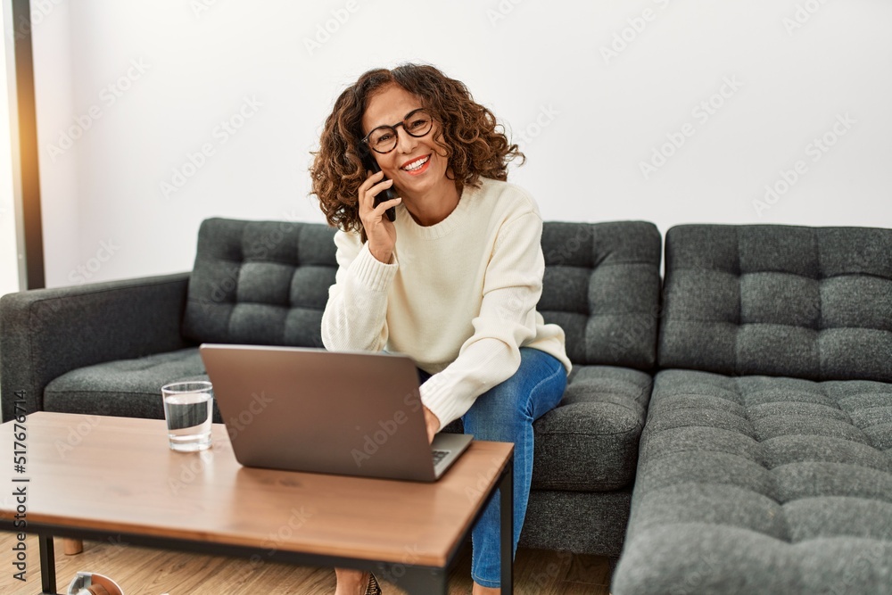 Middle age hispanic woman using laptop talking on the smartphone at home Middle age hispanic woman using laptop talking on the smartphone at home