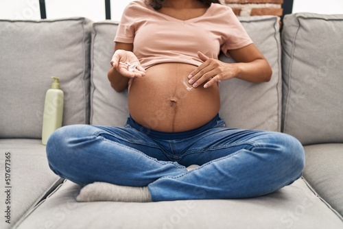 Young latin woman pregnant applying cream lotion on skin belly at home photo