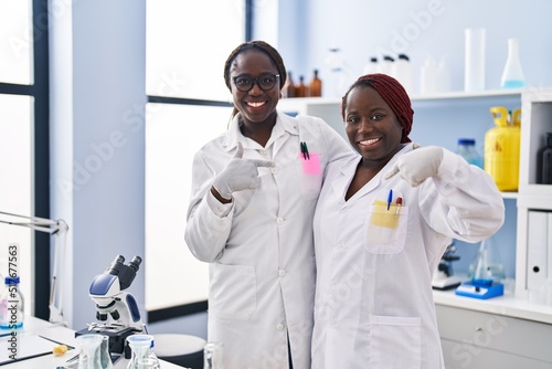 Two african women working at scientist laboratory pointing finger to one self smiling happy and proud