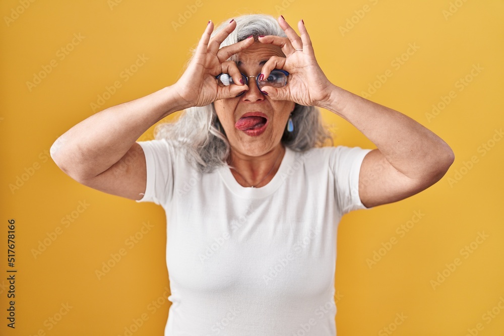 Middle age woman with grey hair standing over yellow background doing ok gesture like binoculars sticking tongue out, eyes looking through fingers. crazy expression. Middle age woman with grey hair standing over yellow background doing ok gesture like binoculars sticking tongue out, eyes looking through fingers. crazy expression.