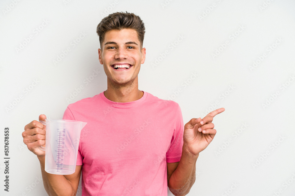 Young caucasian man holding measuring jug isolated on white background smiling and pointing aside, showing something at blank space. Young caucasian man holding measuring jug isolated on white background smiling and pointing aside, showing something at blank space.