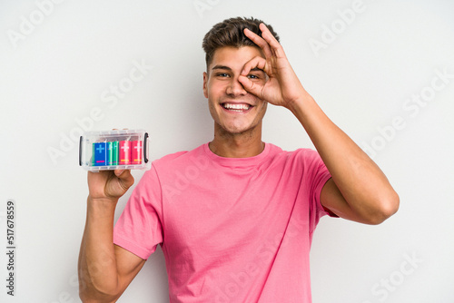 Young caucasian man holding a batteries box isolated on white background excited keeping ok gesture on eye. © Asier