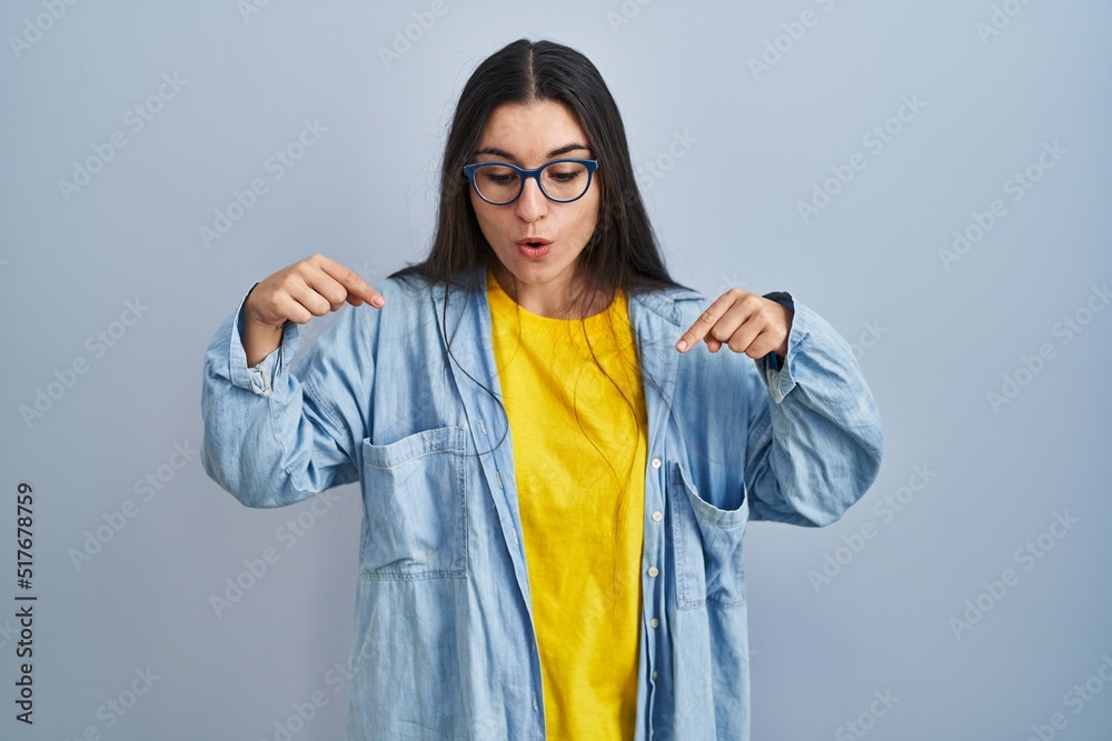 Young hispanic woman standing over blue background pointing down with fingers showing advertisement, surprised face and open mouth Young hispanic woman standing over blue background pointing down with fingers showing advertisement, surprised face and open mouth